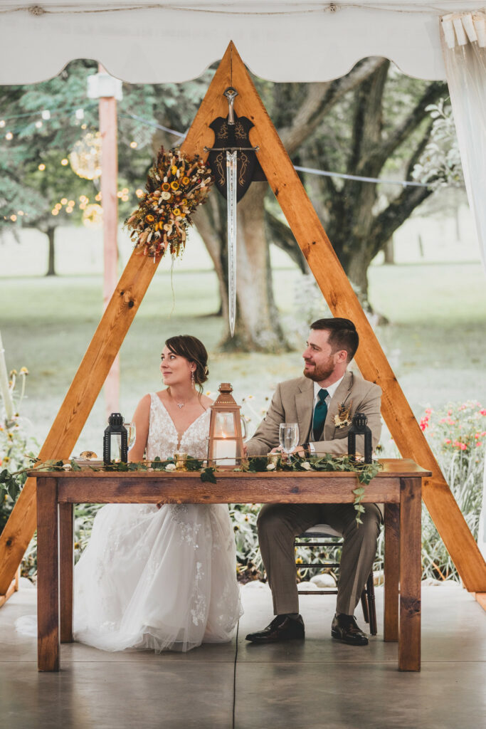 Newlyweds sitting at their sweetheart table under sword and floral.