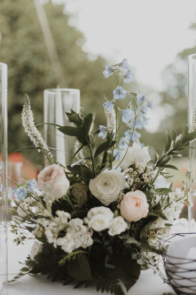 Table centerpiece, summer florals and lush greens with tall candles inside glass hurricanes.