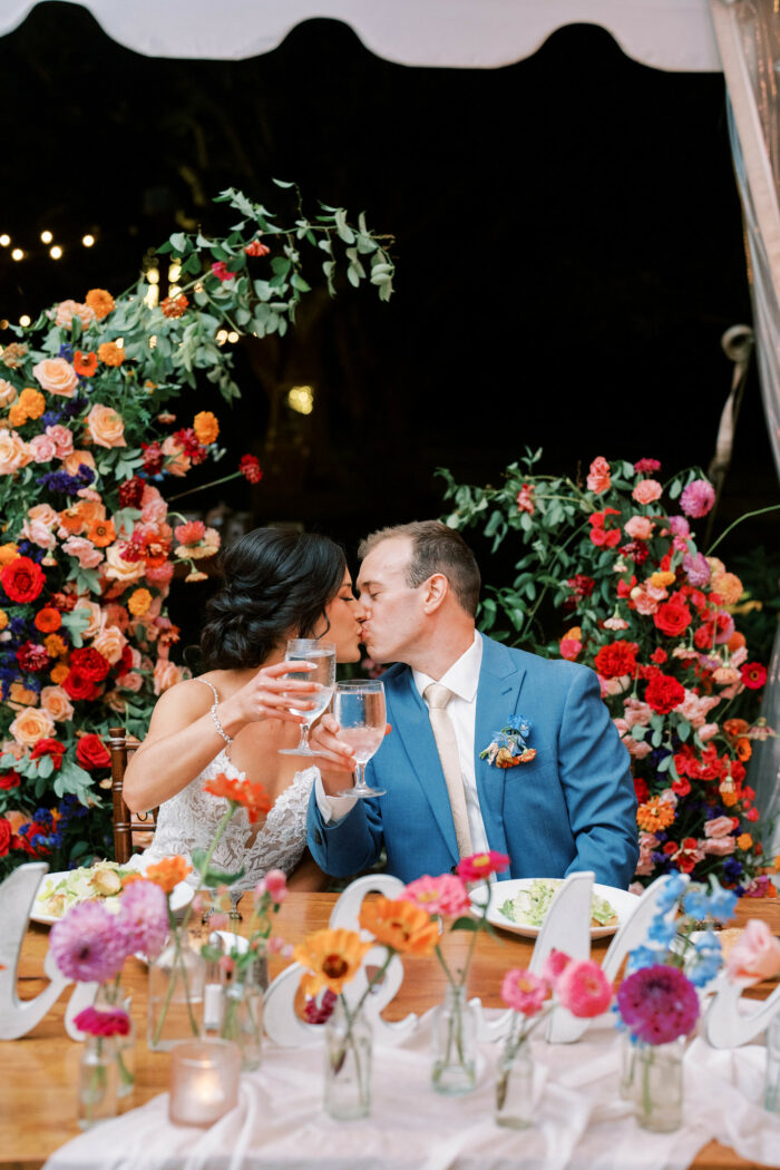 Newlyweds kiss at their sweetheart table surrounded by lush flowers.