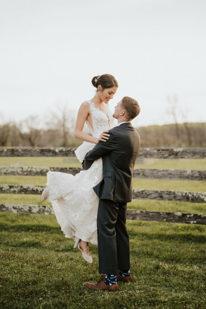 Groom picking up bride in front of farm fence.