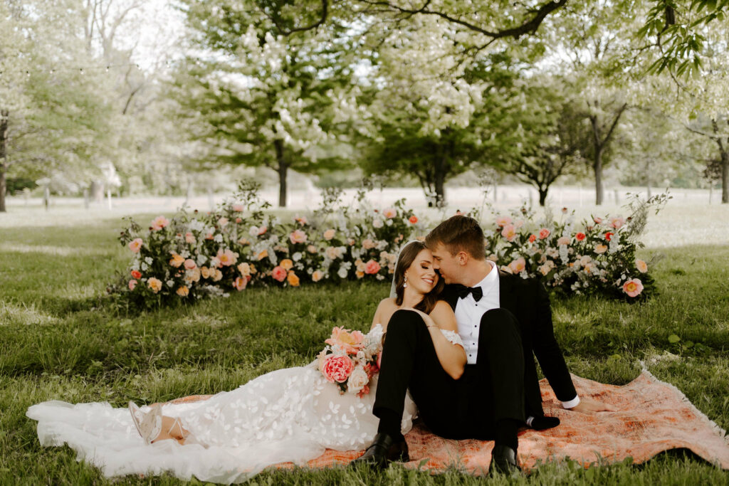 Newlyweds laying on rug on the grass in front of lush flowers.