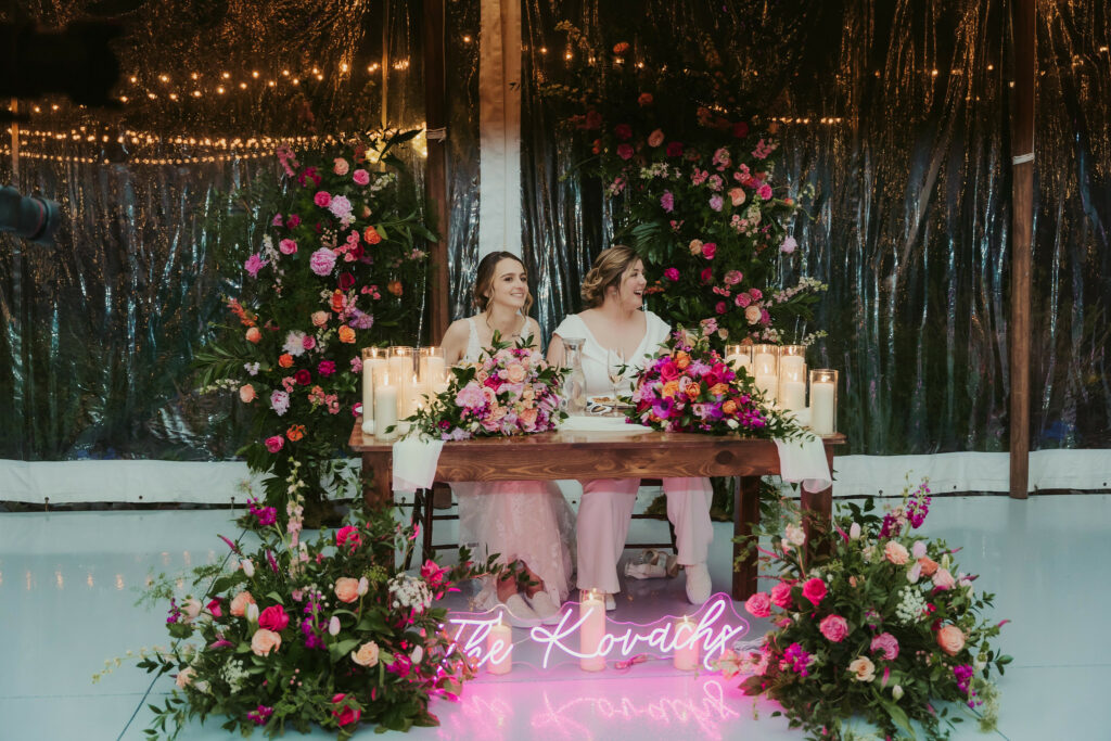 Newlyweds at head table surrounded by a floral decor and glowing neon sign.