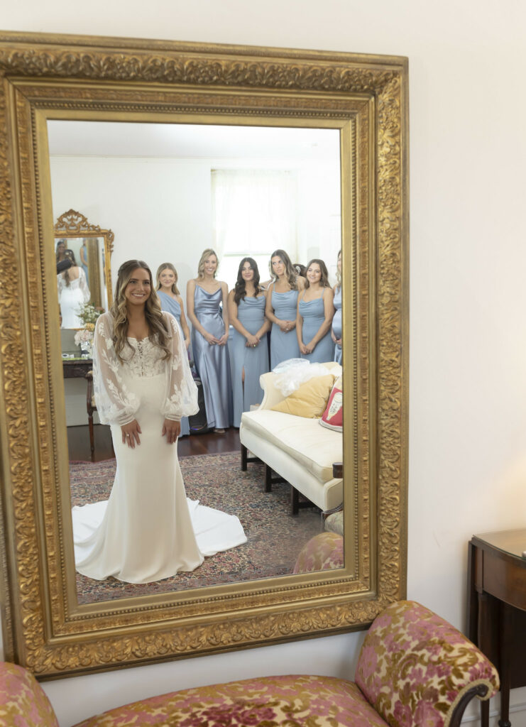 Bride posing in front of a large mirror with bride maids behind her.